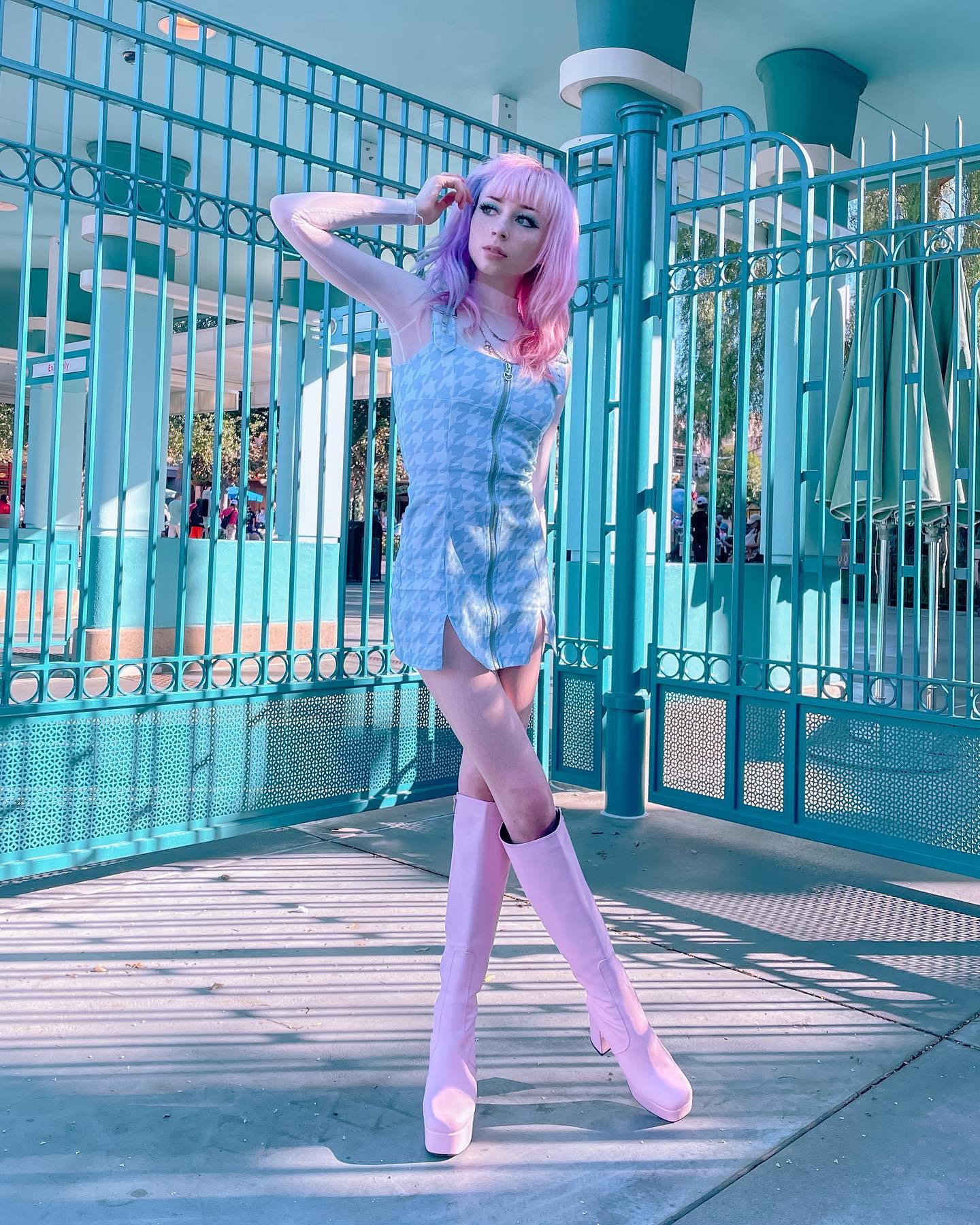 a young woman with pastel pink and purple hair is posing outdoors in front of a teal metal gate. she is wearing a light blue ginghampatterned mini dress with a zipper front and a pair of pastel pink gogo boots with platforms.  she is stylishly dressed in a retro/vintage style, and the overall aesthetic is light, airy, and pasteltoned.  the setting appears to be a park or public space.