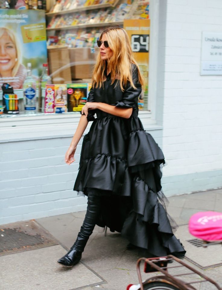 a woman is walking down the street wearing a black dress with ruffles, black leather pants or leggings, and black ankle boots. she is also wearing sunglasses. the woman is walking in front of a store with a window display. a bicycle is partially visible in the lower right corner of the image.