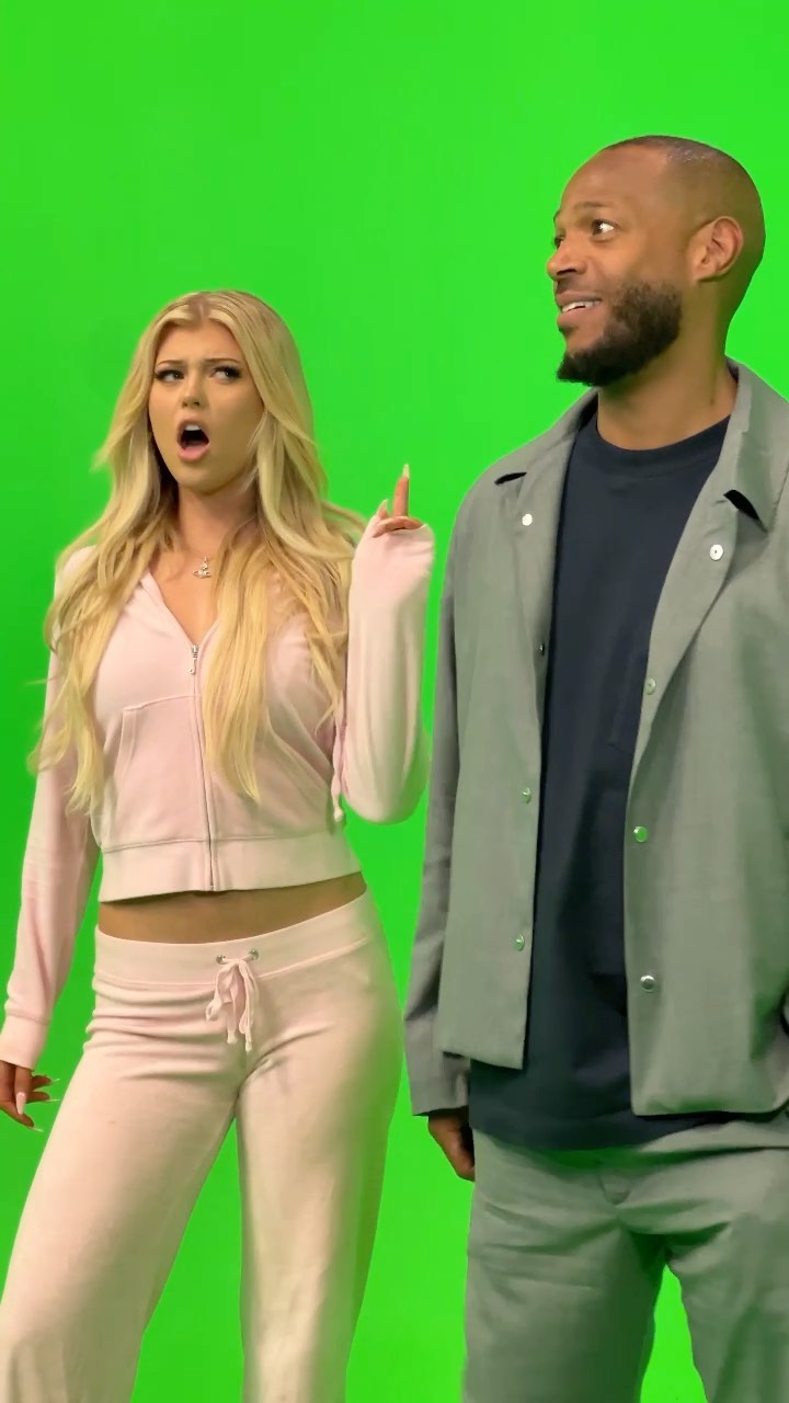 a woman in a pink tracksuit points with her mouth open in surprise while standing next to a man in a jacket against a green screen. the woman is blonde and wearing a crop top. the man is bald and has a beard.