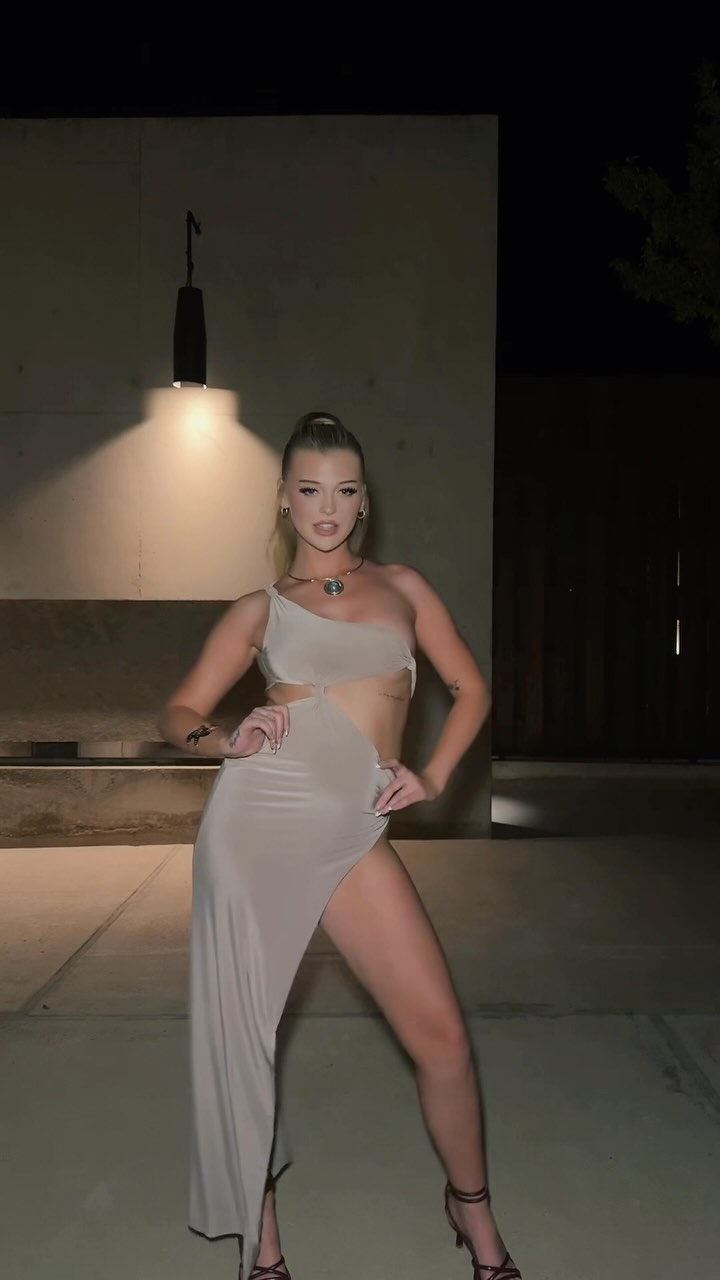 a blonde woman is posing outdoors at night. she is wearing a beige oneshoulder dress with a high slit and high heels. she is also wearing jewelry, including a necklace. she has a confident expression on her face. a street light is shining on her.