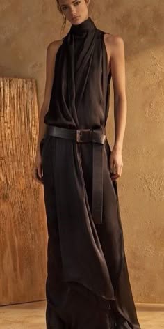 a woman is posing for the camera in a long black dress, the dress has a high neck and is belted at the waist. she has a dark, elegant style and appears to be in a studio setting.