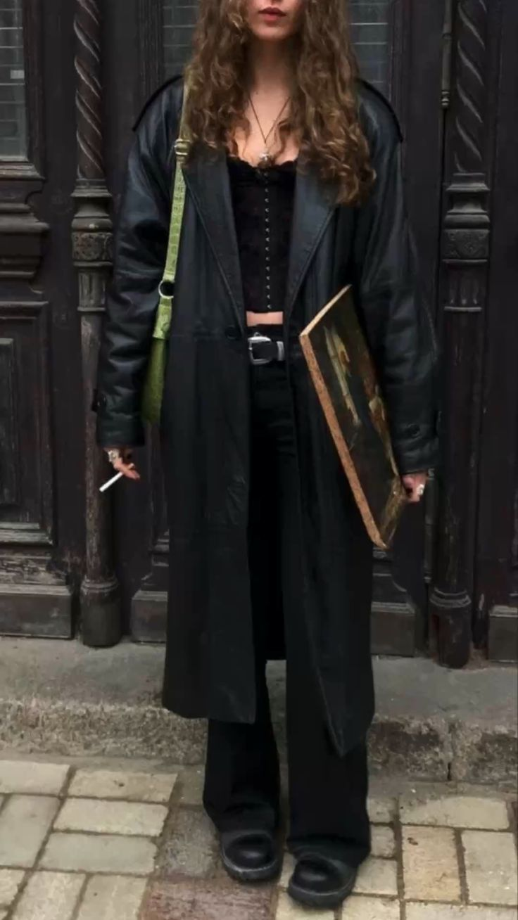 a young woman wearing a black leather coat, black pants, and black boots, holds a framed painting and smokes a cigarette while standing on a city street. her dark hair falls to her shoulders, and she appears to be looking at something offcamera to the right.