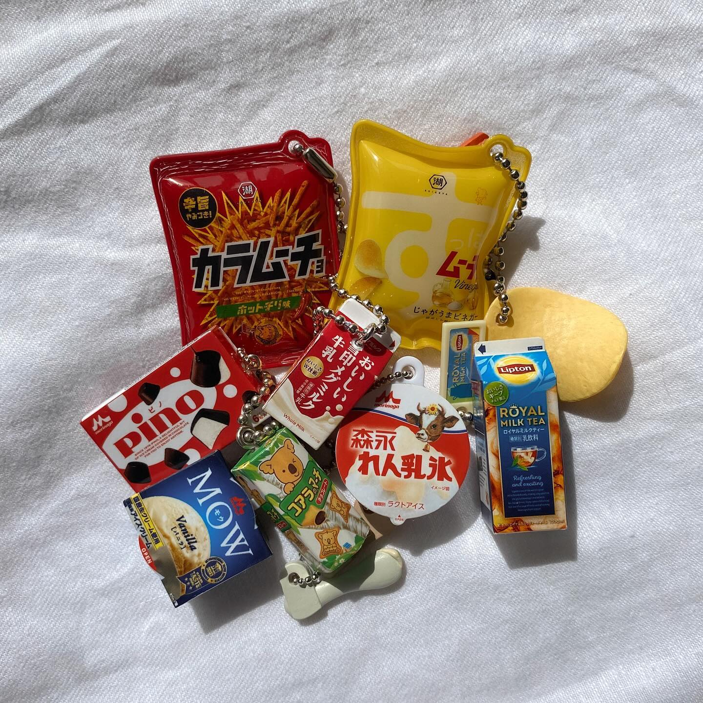 the image shows a collection of japanese snack and candy keychains arranged on a white wrinkled surface. the keychains include miniature versions of popular japanese snack products, such as karamucho chips, muji chips, pino candies, mow ice cream, koala's march cookies, lipton royal milk tea, morinaga milk, and ice cream. they are all connected with chains and keyrings, indicating they are keychains or charms.