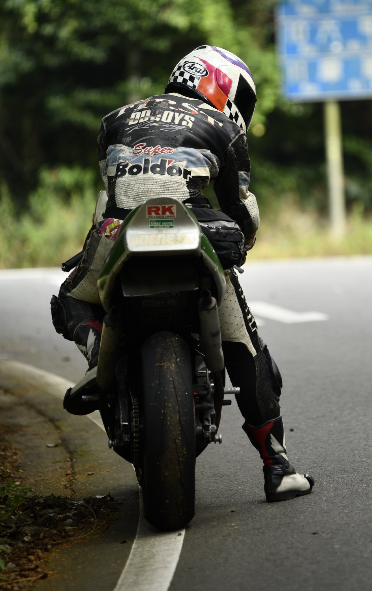 a biker is riding on a paved road on a motorcycle, the biker is wearing a helmet and leather jacket and black and red leather pants. the motorcycle has a green and silver frame, black seat and a black rear tire. the rider is wearing a red and black and white helmet. the rider is looking ahead while riding, the rider has black gloves on their hands.  the image is taken during the day in a bright light.