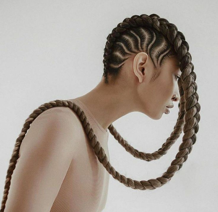 a woman is photographed from the side, looking to the left. her hair is styled in two large braids that loop around her neck. the braids are elaborately designed and have an intricate woven pattern. the woman is wearing a beige top. the image has a minimal aesthetic, with the background being white.  the hair is a statement piece of this portrait.