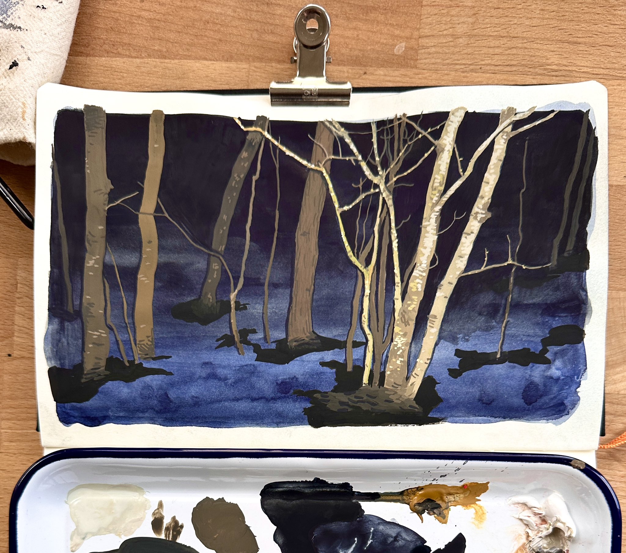 a watercolor painting depicts a dark forest or swamp scene. several trees stand in the water, with some appearing bare. the background is dark and the foreground is a blue watery surface. the painting is clipped to a sketchbook, and below it is a paint palette with various colors.