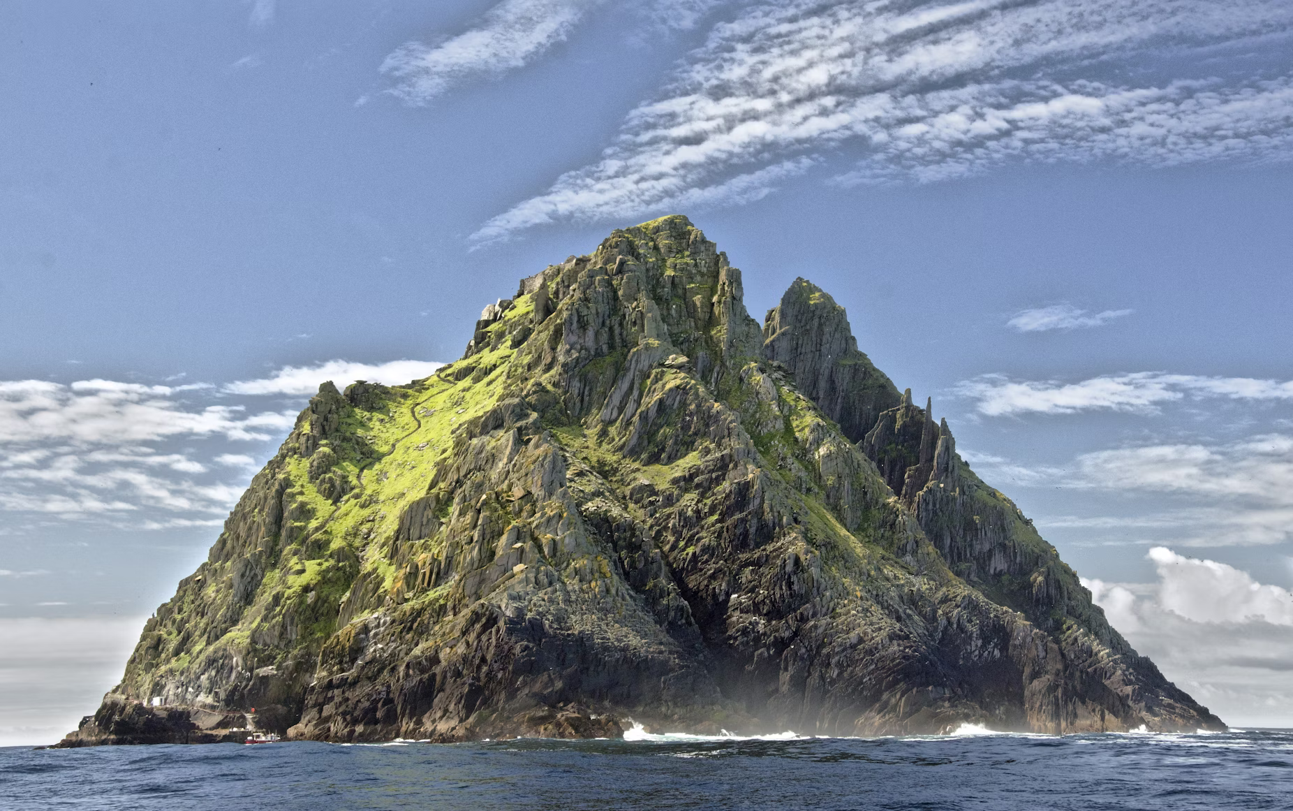 an island mountain rises out of a rough blue ocean with a blue sky and puffy clouds above. the mountainside is green and brown with vegetation and rock.