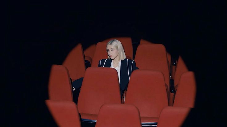 a blondehaired woman in a black blazer and white shirt sits alone in a dark movie theater.  the red seats surround her, and a spotlight highlights her from the front. the overall mood is pensive and mysterious.  her style is sharp and fashionforward. the image could be a still from a music video or a promotional photo shoot for a kpop artist.