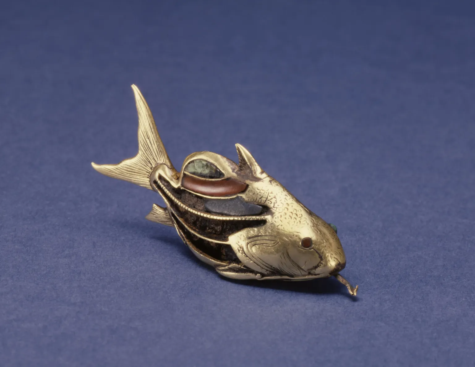 the image shows a gold fish ornament on a blue background. the fish is made of gold and decorated with colorful stones. the fish has a long tail and a small mouth. the fish is lying on its side.