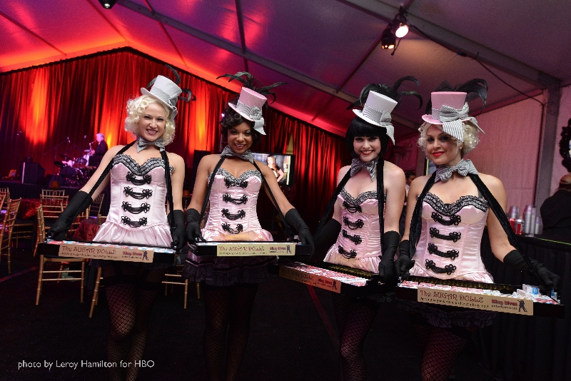 four women dressed in pink burlesque costumes, complete with hats and feathers, are seen serving trays at an elegant event.  they are wearing black gloves and are positioned against a backdrop of red curtains and sophisticated lighting. the image is credited to photographer leroy hamilton for hbo, suggesting the event may be a highprofile one. the overall style evokes a vintage or retro feel, reminiscent of the 1920s or 1930s, with the costumes and setting creating a cabaret or showgirl atmosphere.