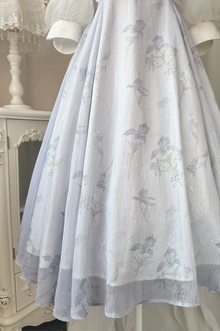 a closeup image of a flowing, white skirt with a floral print. the skirt is made of a delicate, sheer fabric and has a tiered, aline silhouette. the floral print is subtle and delicate, featuring small, light blue flowers scattered across the fabric. the background of the image is a soft, creamy white, which complements the ethereal and romantic feel of the skirt.