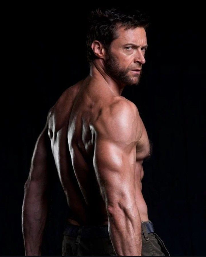 a shirtless muscular man with a toned body and abs is posing in a studio. he has a serious expression on his face and is looking over his shoulder. the background is black.