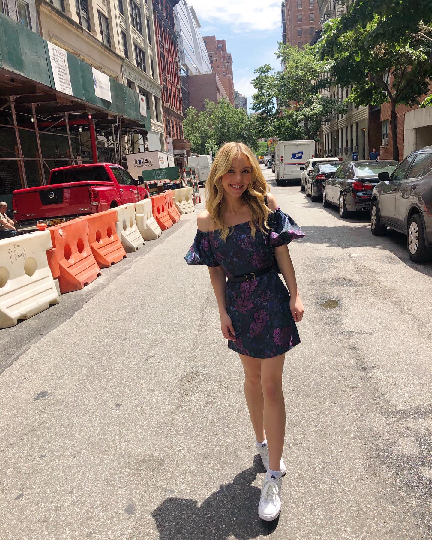 a blonde woman wearing a dark floral offtheshoulder mini dress and white sneakers walks down a city street in new york city.  construction is visible in the background. the sun is shining and it's a summer day.  the overall style is casual and trendy.