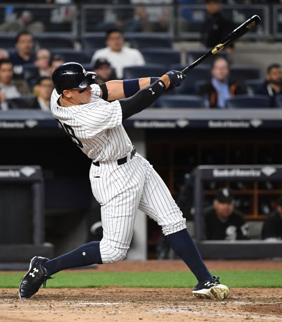 a color image of new york yankees player, aaron judge, swinging a bat during a game. judge is wearing the team's home pinstriped uniform and batting helmet. the background is a blurred view of the stadium and crowd.