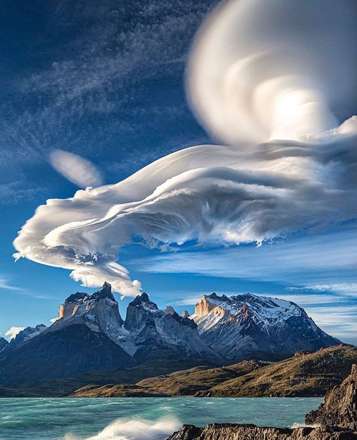 a breathtaking panoramic view of the majestic torres del paine national park in chile. the image captures the rugged beauty of the andes mountains, with towering peaks and snowcapped summits rising dramatically against a backdrop of a vibrant blue sky. a mesmerizing cloud formation, resembling a giant swirl, dominates the upper portion of the image, adding an ethereal touch to the scene. the clear blue sky is punctuated by wispy clouds, further enhancing the dramatic nature of the landscape. a tranquil lake stretches out in the foreground, reflecting the surrounding mountains and the azure sky, creating a serene and idyllic atmosphere. the image showcases the aweinspiring power and beauty of nature, inviting viewers to immerse themselves in the tranquility and grandeur of this remote and pristine wilderness.