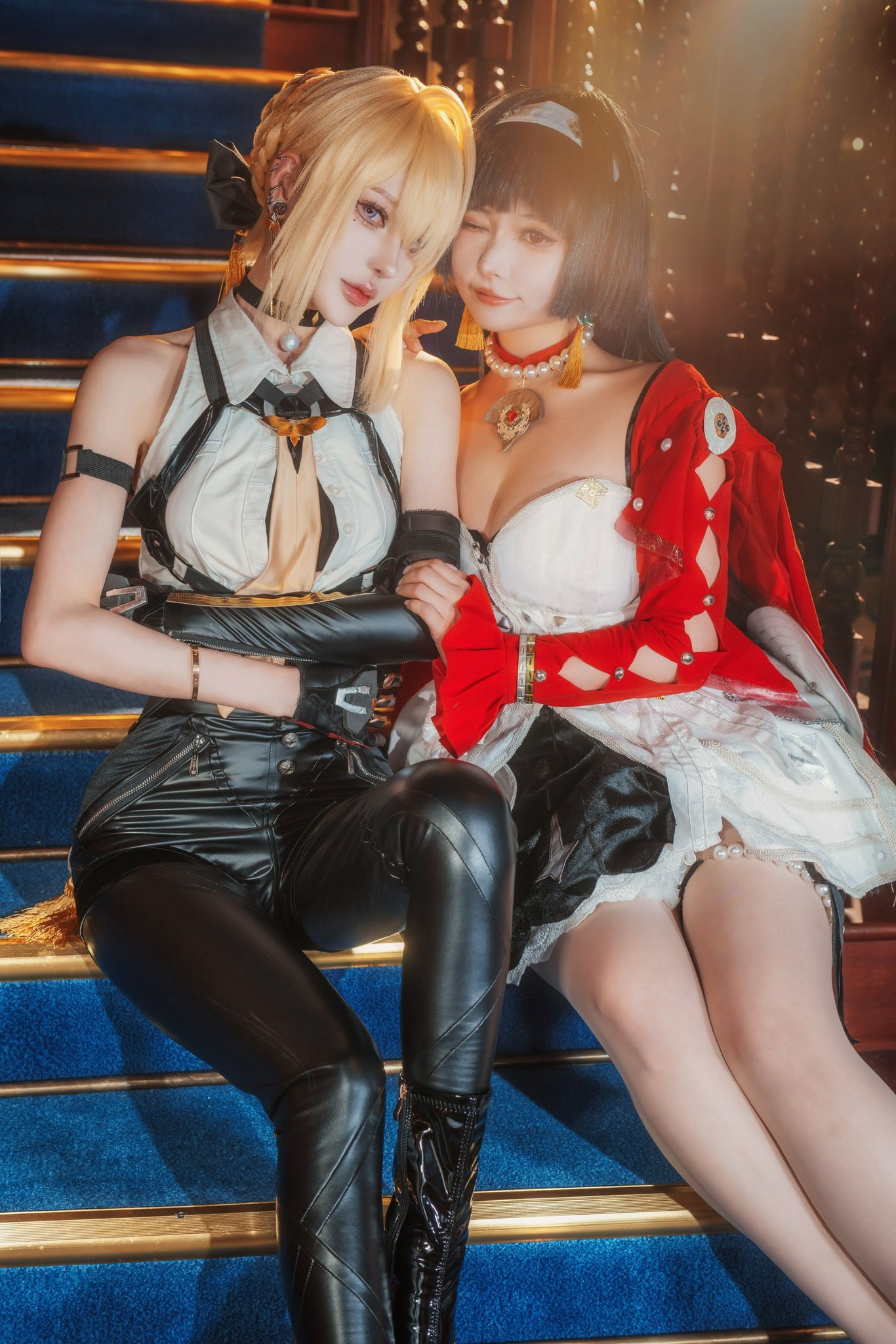 two women in elaborate cosplay costumes are sitting on a staircase.  one is cosplaying a character with blonde hair and a black leather outfit. the other is cosplaying a character with dark hair, and is wearing a white and red dress with black accents. they are sitting close together, appearing friendly and possibly portraying characters from the same fictional universe. the setting is indoors, on a plush, dark blue staircase with gold accents.  the image is well lit, sharply focused, and exhibits a professional photography style.