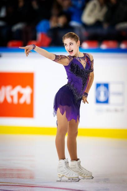 a young female figure skater in a purple dress performs on an ice rink. she is smiling and extending her right arm out toward the left of the frame.  her left hand is resting on her hip. she is wearing white skates and is on the ice.  the image is taken from a low angle. the ice rink is a light color and the background is blurred.  the skater is in midperformance.  the overall image suggests a feeling of excitement and grace.