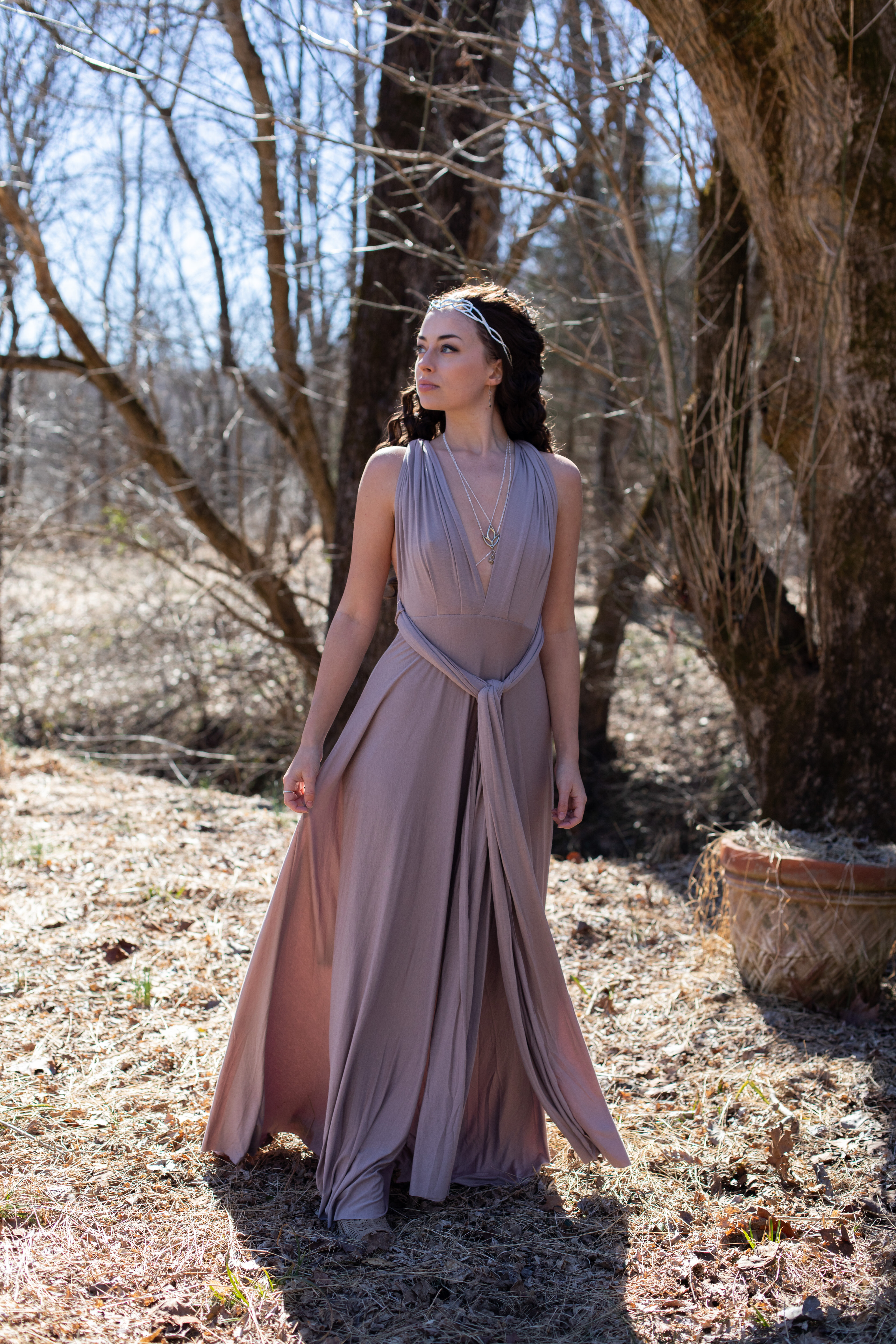 a young woman in a flowing mauve dress stands in a sundappled forest. she is looking off to the side, her dark hair loose around her shoulders. the dress has a halter neckline and a tie at the waist. she is wearing a delicate silver headband and necklace. the forest floor is covered in dry leaves and there is a woven basket in the background. the overall feeling of the image is ethereal and mystical.