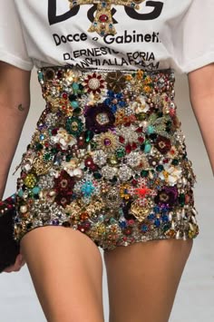 a woman is wearing a white tshirt with a sparkling, bejeweled, miniskirt/short. the shirt has a black graphic design and says "docce & gabinetti" on the front. she has on a black clutch purse on her left arm. she is standing and posing. the setting is not clear but it looks like the woman is on a white backdrop with a blurred background. the focus of the image is on the dazzling skirt/short.