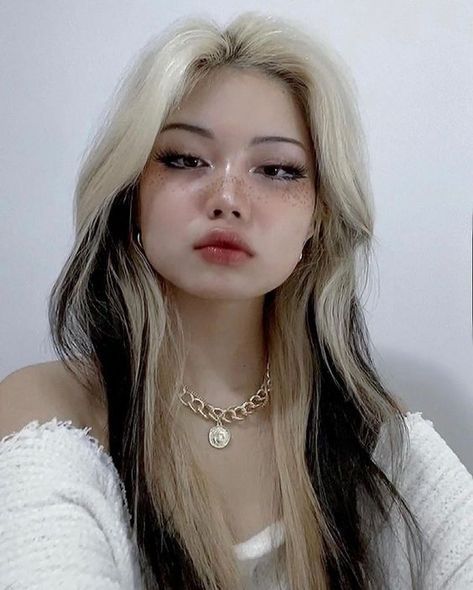 a young woman with a trendy twotoned hairstyle featuring blonde and black streaks poses for a portrait against a minimalist white background. she wears naturallooking makeup that accentuates her features and a chunky gold chain necklace that adds a touch of edge to her look. the soft lighting enhances her features and creates a dreamy atmosphere.
