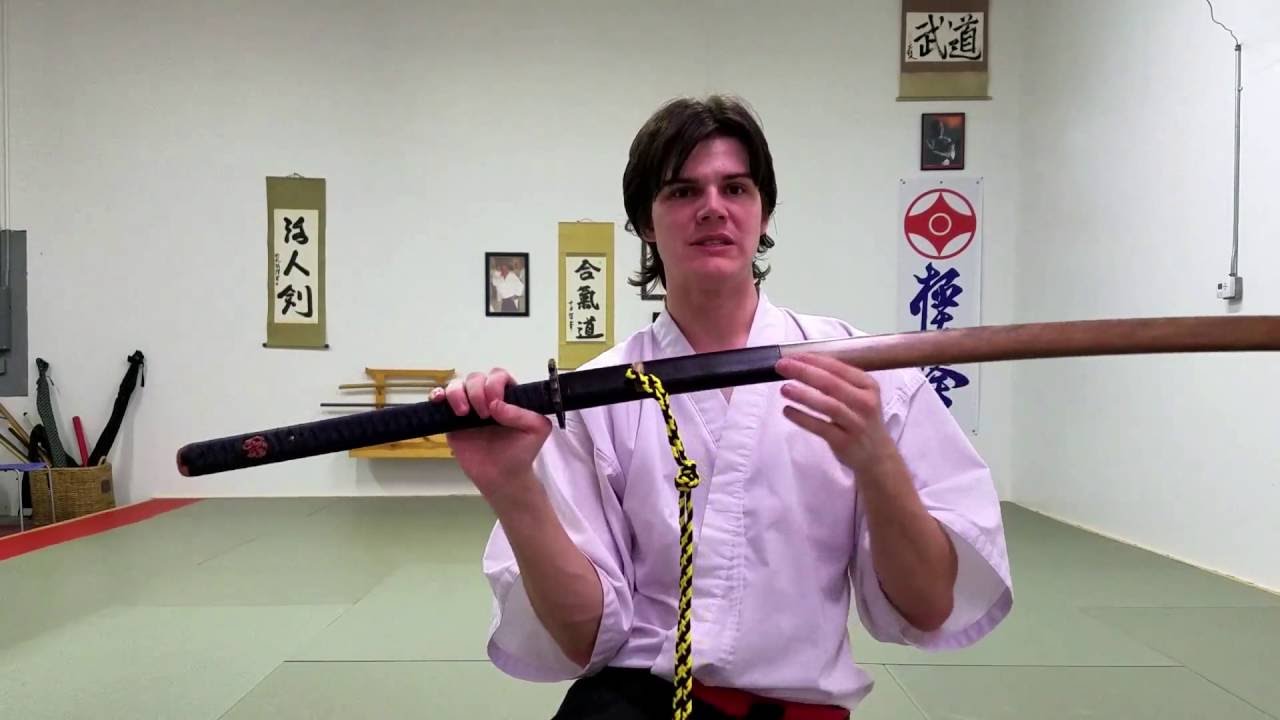 a man wearing a martial arts uniform is holding a katana, which is partially drawn from its scabbard. the man is in a dojo, a training space for martial arts. the uniform includes a white jacket and a black hakama, with a red obi around the waist. the katana has a black scabbard with a sageo cord. the sword's wooden blade is visible. there are japanese scrolls and other martial arts paraphernalia in the background.
