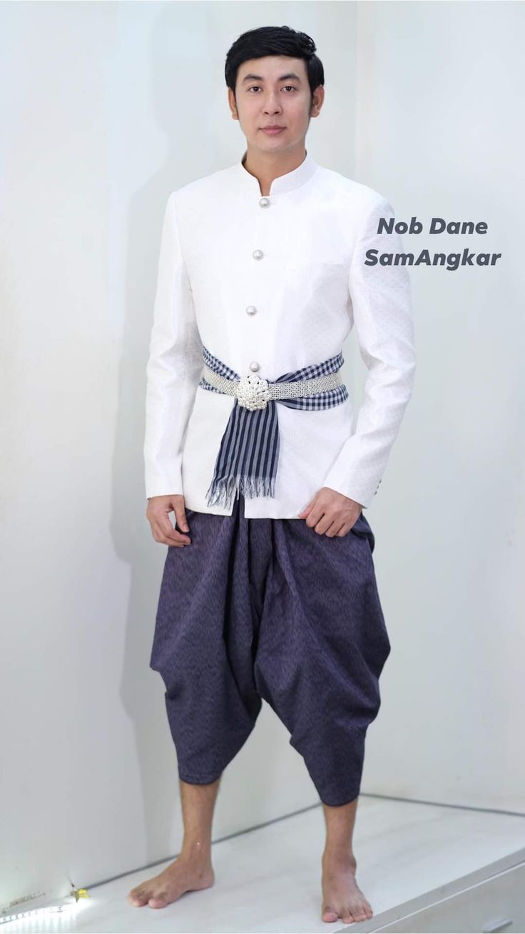 a man is standing in a studio, wearing traditional thai clothing. he is smiling and looking at the camera. he is wearing a white shirt with a sash around his waist. he is wearing blue pants. the background is white.