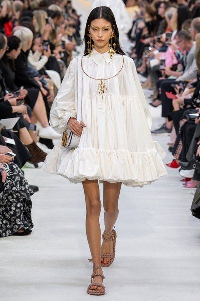 a young model wearing a white dress and sandals walks the runway during a fashion show. she has a beautiful white dress with a ruffled hem and big sleeves. the model is wearing accessorized with a delicate gold necklace, and matching golden earrings.  her hair is pulled back into a sleek ponytail with a middle part and she is wearing light makeup. the model looks confident and elegant as she struts down the runway.
