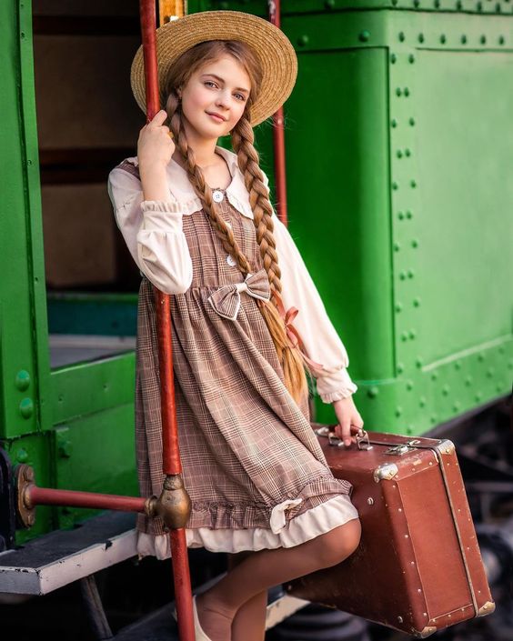 a young girl in a vintageinspired dress and straw hat stands on the steps of a green train, holding a brown suitcase. her long blonde hair is braided and she looks directly at the camera.