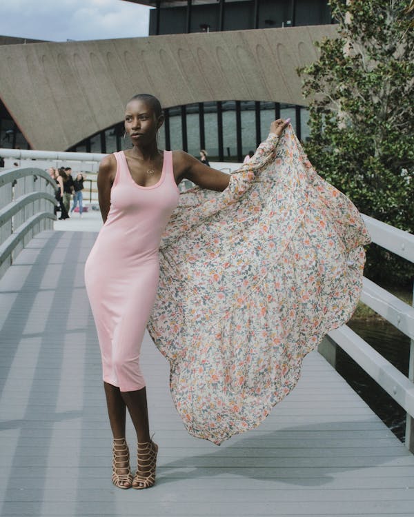 the image shows a stylish young woman with short, dark hair and dark skin standing on a modern pedestrian bridge.  she is wearing a light pink midi dress and a floral patterned shawl or scarf draped over her shoulders.  she is posing confidently, wearing stylish heeled sandals. the background features contemporary architecture and lush greenery. the overall feel is chic and modern.