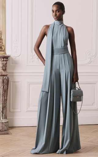 a woman in a light blue formal outfit stands against a white wall and looks at the camera.  she is wearing a halter top with a pleated skirt, and she is carrying a white handbag.  her outfit is very stylish and sophisticated, and she is looking elegant.