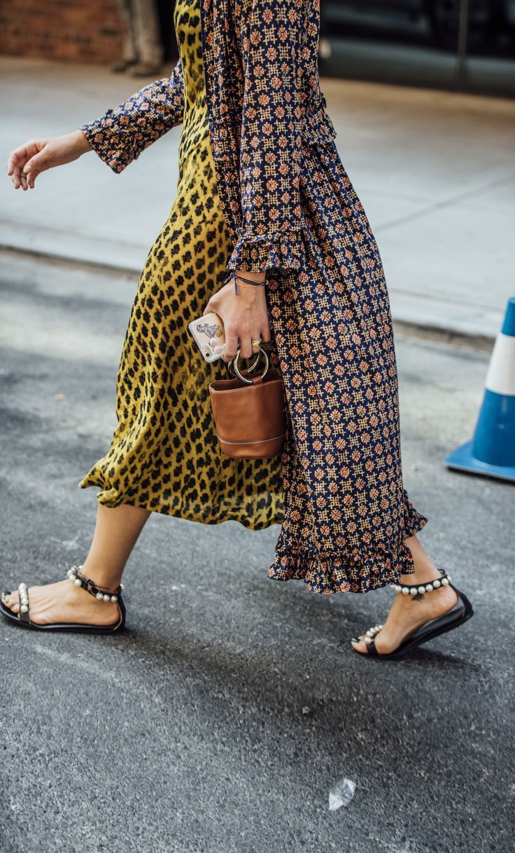 a woman is walking down a city street wearing a long, printed dress. the dress is a mix of yellow and dark colors with a geometric pattern. she is carrying a small leather bucket bag and wearing flat sandals with pearl details. her style could be described as bohemian or eclectic. the photo is taken from a slightly low angle, showing the woman in motion. the background is blurred, with a glimpse of a brick wall and a traffic cone visible.