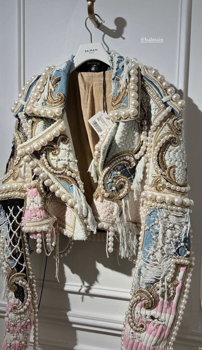 an elaborate balmain jacket hangs on a hanger. the jacket is heavily embellished with pearls and other decorative elements. the jacket has a textured, possibly tweed or woven, base, and the pearls are arranged along the edges and seams. the garment is clearly a designer piece and is being showcased as such.