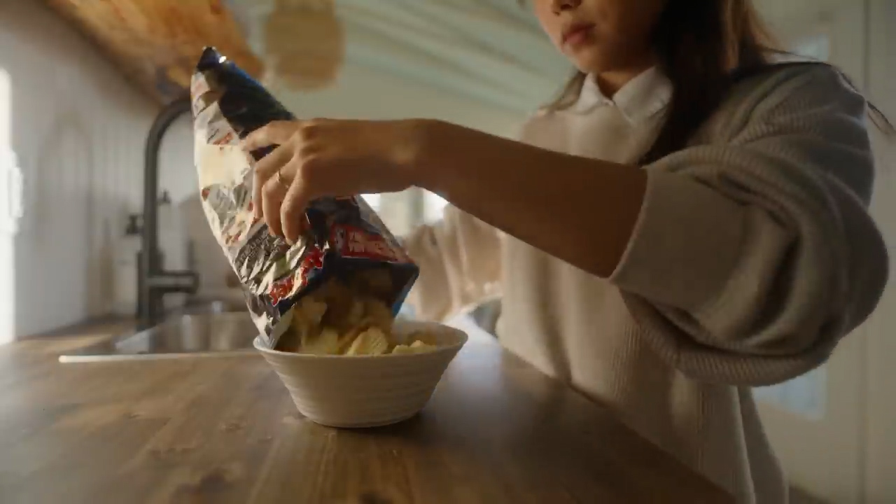 a young girl is pouring a bag of potato chips into a white bowl. the scene takes place in a kitchen, with warm, natural light illuminating the countertop and the girl's hands. the focus is on the act of pouring, highlighting the chips and the bowl.
