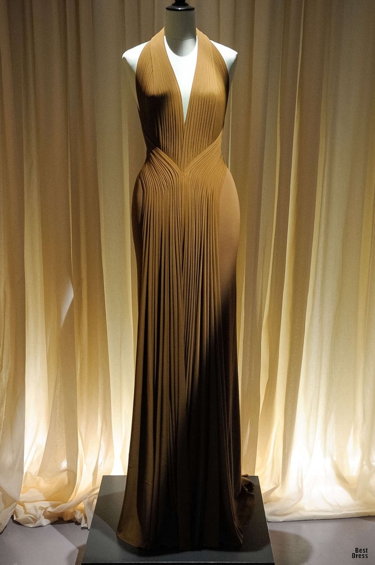 a fulllength studio shot features a brown, floorlength halter neck gown displayed on a mannequin against a backdrop of shimmering beige curtains. the dress boasts an elegant design with intricate pleating details, especially in the bodice area which flares out towards the waist. the silhouette is fitted, emphasizing the mannequin's form, before cascading down into a graceful, flowing skirt. the mannequin stands on a square, gray platform that elevates the garment, enhancing its presentation. the lighting is soft, highlighting the texture and sheen of the dress's fabric and the curtains, creating a luxurious and refined aesthetic. the phrase "best dress" is subtly visible in the lower right corner.