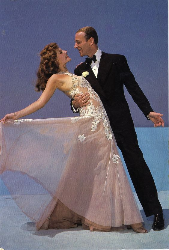 fred astaire and ginger rogers in a dance pose on a stage, bathed in theatrical lighting. ginger rogers wears a flowing peach gown with floral appliqués, while fred astaire is in a black tuxedo. their dynamic pose, with rogers leaning back, captures the energy and grace of their iconic dance partnership.