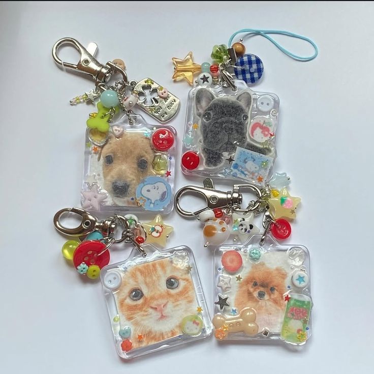 a highangle, closeup shot displays a collection of four squareshaped keychains on a plain white surface. each keychain features a clear resin casing encasing a printed image of an animal – three dogs and one cat. the dog breeds appear to be a tan mixed breed, a french bulldog, and a pomeranian. the cat is an orange tabby. each keychain is adorned with various colorful beads, charms, and miniature decorative items embedded in the resin, such as stars, buttons, and small characters. a lobster clasp is attached to each keychain, allowing it to be connected to keys or bags. the overall aesthetic is cute, handcrafted, and personalized, likely intended as gifts or novelty items.