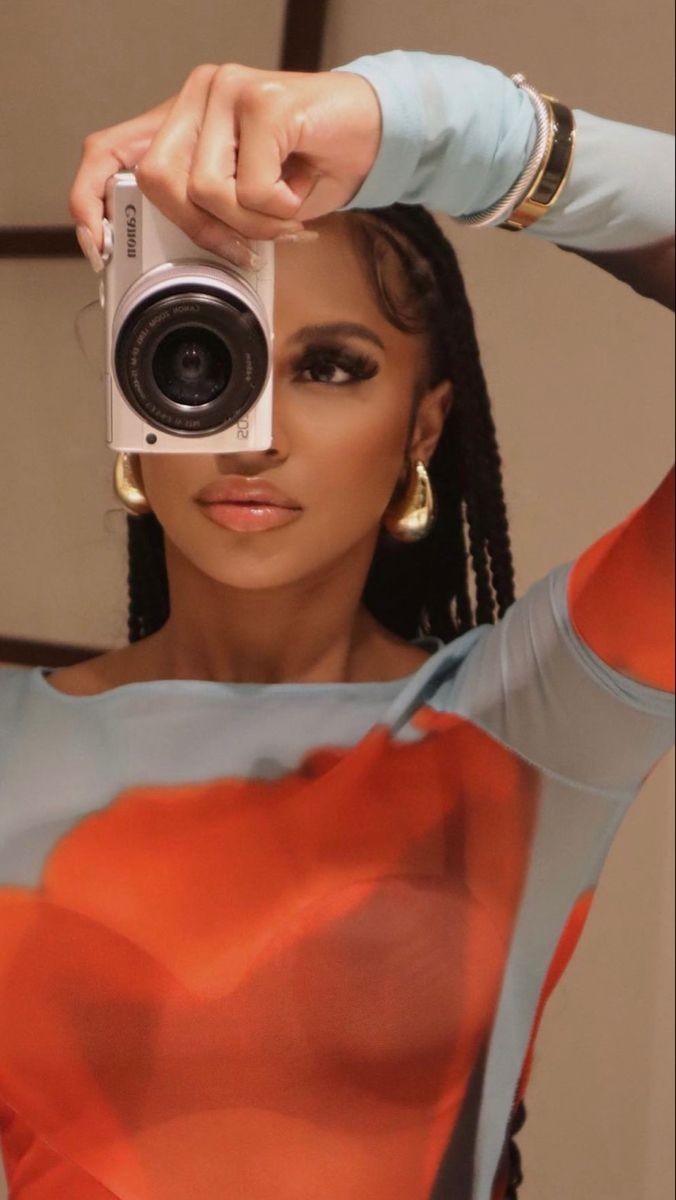 a woman with dark braids and makeup is posing with a canon camera in front of a mirror. she's wearing a sheer blue top with an orange top under it and a gold bracelet. the picture is a classic fashion shoot with a modern look and a contemporary style.