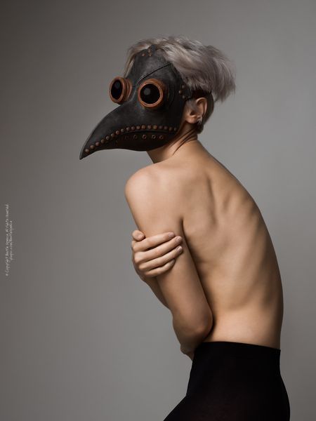 a woman with a plague doctor mask on is standing in a studio. she is wearing black tights and her body is turned to the side. the background is gray.