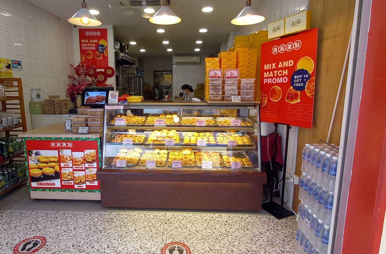 a storefront of a chinese bakery with a display case filled with egg tarts, a sign promoting a mix and match offer on the egg tarts, and a shelf of water bottles.
