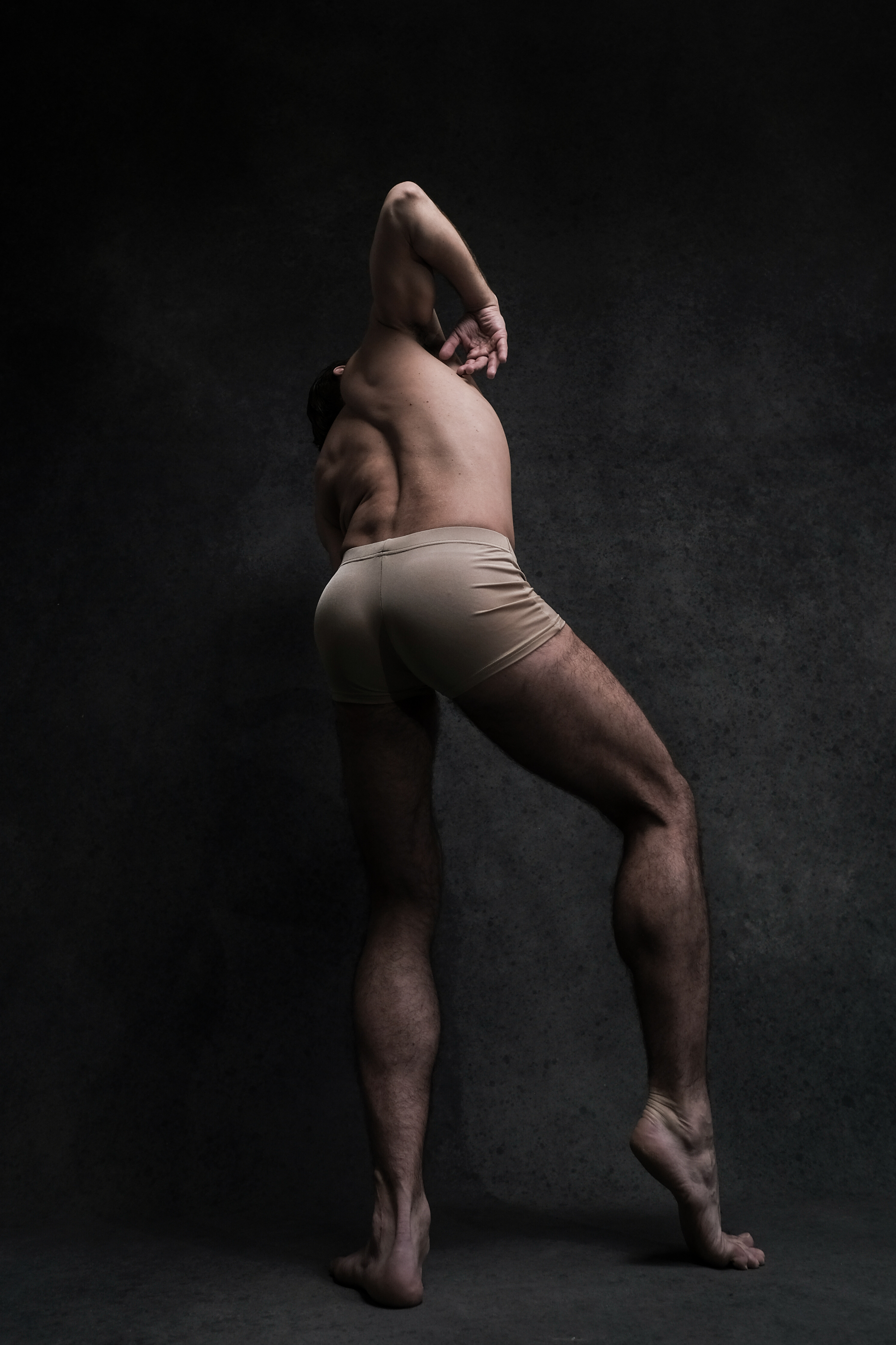 a male dancer in tan underwear is captured midpose against a dark grey backdrop. the perspective is from the rear, showcasing the dancer's muscular back, toned legs, and the elegant arch of his body. he is barefoot, highlighting the raw and natural aesthetic of the image.  the lighting is dramatic, emphasizing the contours of his physique and creating a sense of strength and artistry.