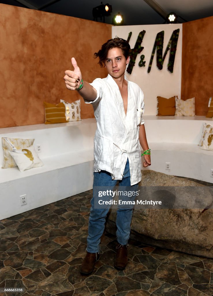 a full shot of cole sprouse, an actor, model, and celebrity, smiling and giving a thumbsup. he is wearing a white shirt, jeans, and brown boots. he is standing in front of an h&m backdrop, with a stone to his right and a bench to his left. the floor is a mosaic of stones.