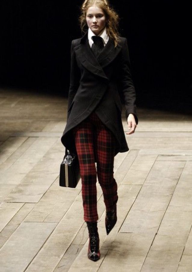 a woman in a black suit and red plaid pants is walking down a runway. she is wearing black boots and a white shirt with a black tie.