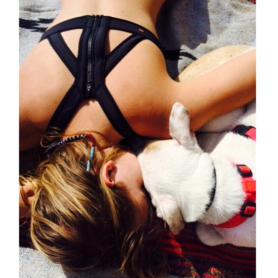 the image shows a young blonde woman lying on a patterned blanket outdoors, sunbathing. she is wearing a black sports bra with a zipper detail. a white french bulldog puppy is nestled close to her, resting its head on her chest and shoulder.  the woman and dog appear relaxed and comfortable, suggesting a scene of summer leisure and affection between a person and their pet. the setting appears to be a sunny beach or similar outdoor location.