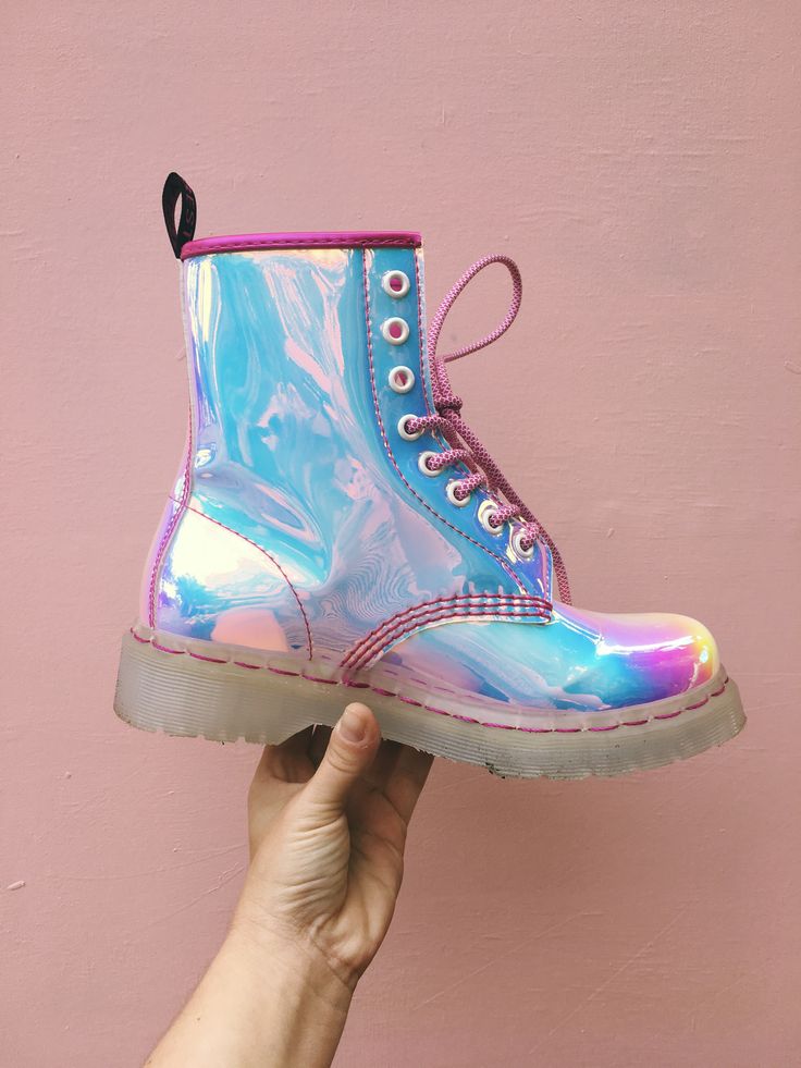 a closeup shot showcases a single iridescent dr. martens 1460 boot. the boot has a holographic, pastel pink, blue, and purple finish.  it features a transparent sole.  a person's hand is gently holding the boot against a light pink wall. the overall feel is fashionable and trendy.
