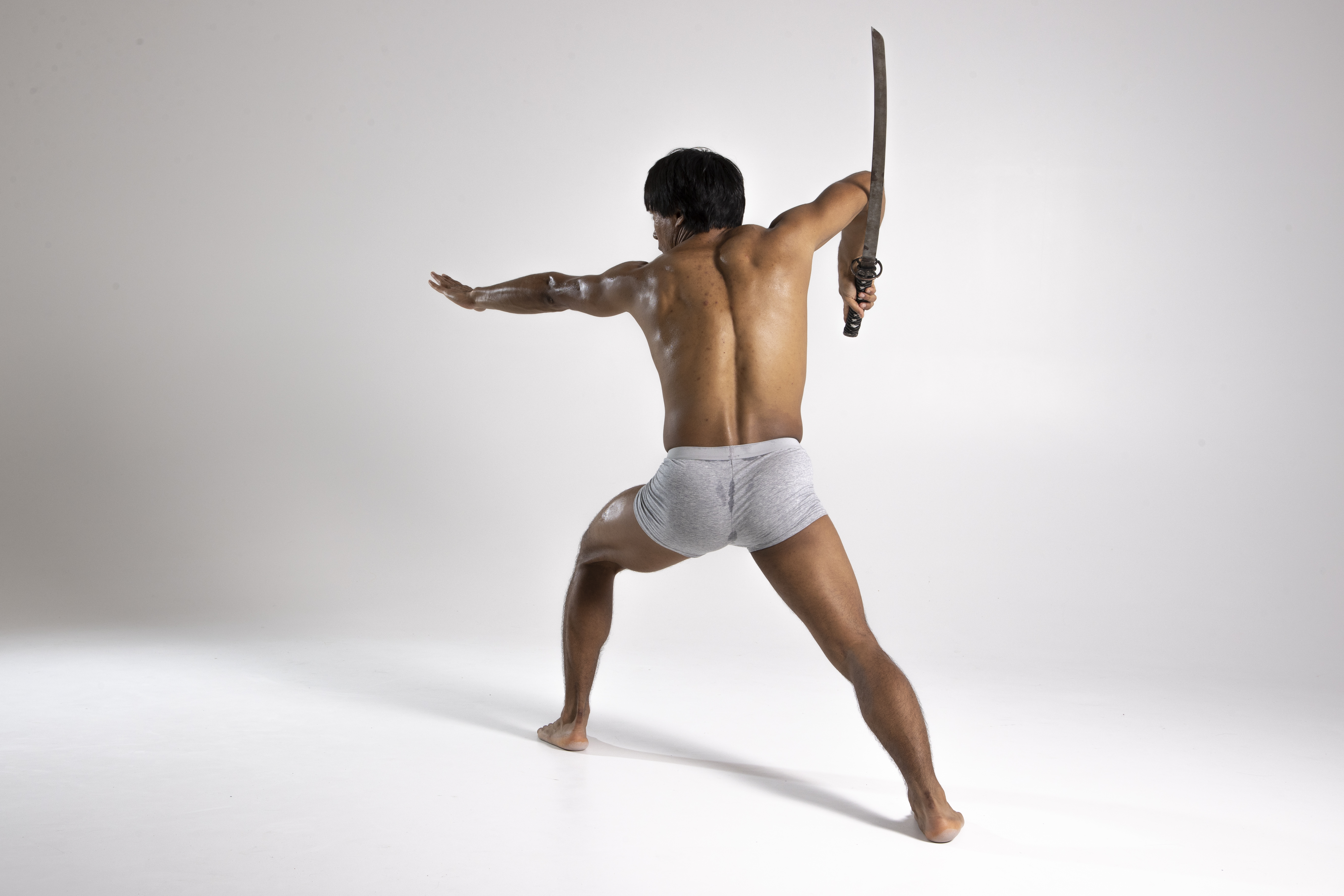 a shirtless asian man in grey underwear is posing with a sword in a martial arts stance. he is viewed from the back, with his right arm holding the sword above his shoulder and his left arm extended to the side. he has a muscular build and his posture conveys strength and readiness. he is positioned on a white floor with a white background.