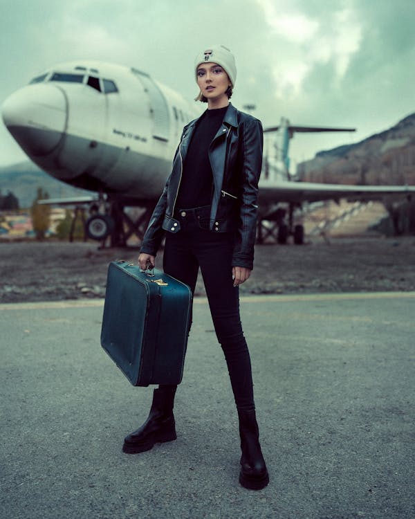 a fulllength photo of a young woman standing on an airport tarmac in front of a large, seemingly abandoned, vintage airplane. she is carrying a dark teal vintage suitcase. she is dressed in a black leather jacket, black skinny jeans, black ankle boots, and a white beanie. her expression is serious and somewhat pensive. the overall mood of the image is moody and atmospheric, with an overcast sky providing a dramatic backdrop. the setting and attire suggest a sense of travel and perhaps departure or arrival.