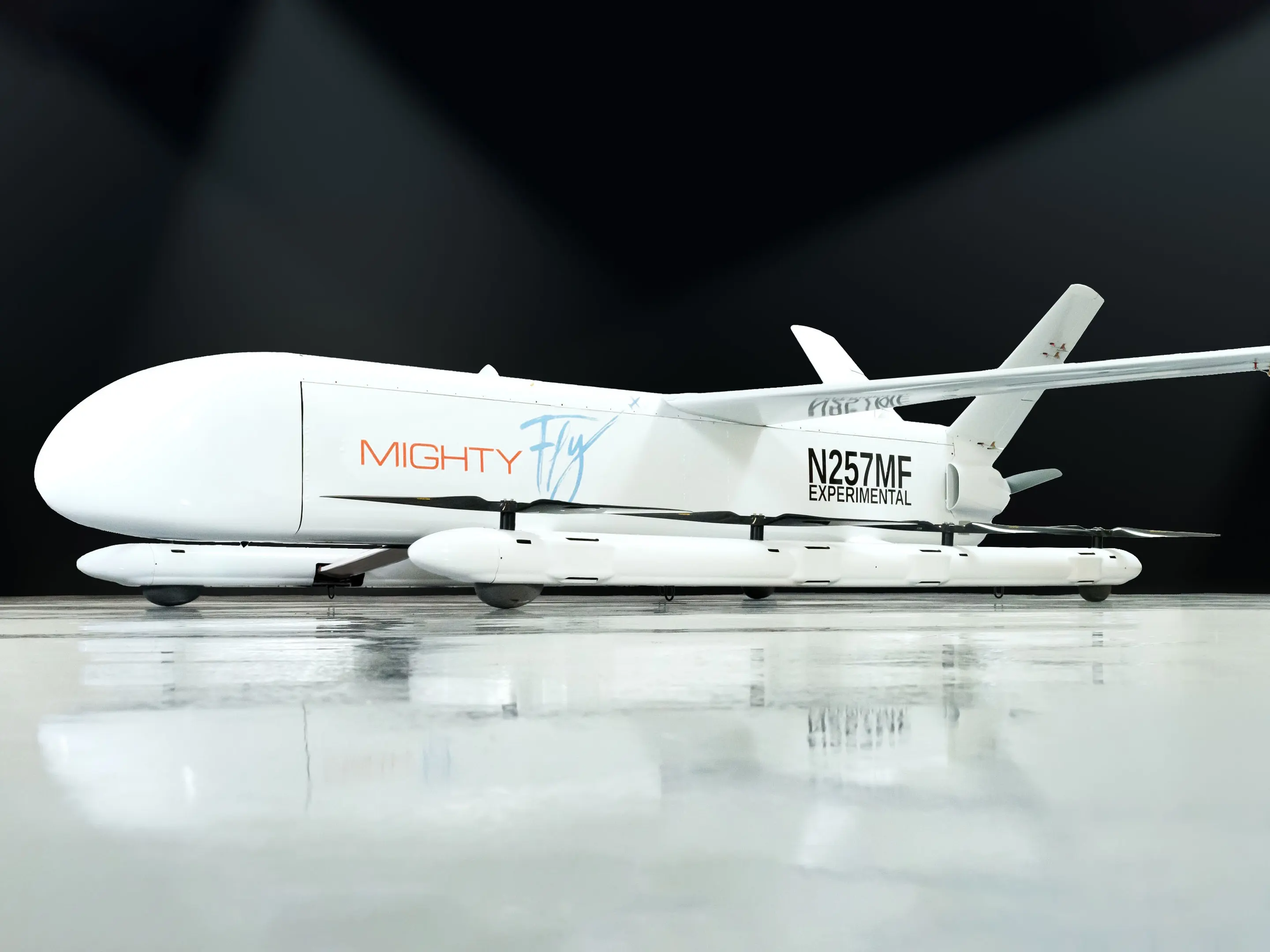 a white airplane with the words "mighty fly" on the side and a black "n257mf experimental" in the back.  it is sitting on a gray floor and appears to have a lot of lift from the rear,  giving the illusion of a flying car.