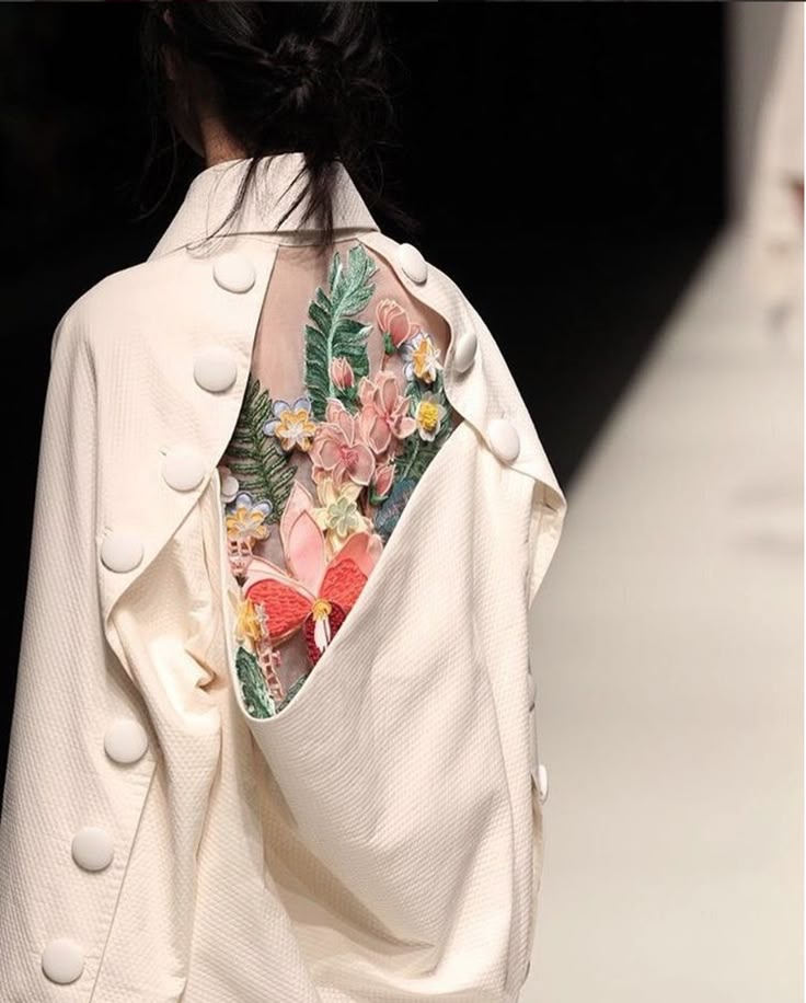 the image shows the back of a white jacket worn by a woman with a floral design. the jacket has large, round white buttons running down the back and along the shoulders. there is a panel of sheer fabric in the middle of the back revealing embroidered flowers and leaves, including a butterfly. the texture of the white fabric is visible, and the setting appears to be a runway show. the woman's hair is styled in a messy bun.