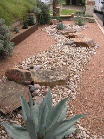 this image showcases a beautifully designed xeriscape garden.  a curving pathway of reddish gravel is accented with various sizes of river rocks and larger natural stones.  the pathway is punctuated by strategically placed droughttolerant plants, most notably a large agave plant towards the bottom. other smaller succulents and desert grasses are visible.  the overall effect is a visually appealing, lowmaintenance landscape ideal for arid or semiarid climates. a rustycolored metal planter box with plants is visible in the upper left.  the background includes a portion of a house and a street.