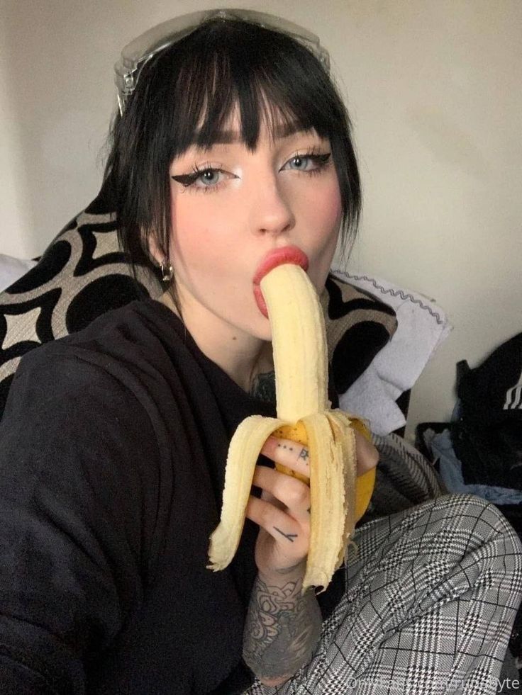 a woman with dark hair and eye makeup is eating a banana while sitting indoors. she is wearing plaid pants and has tattoos on her arm.
